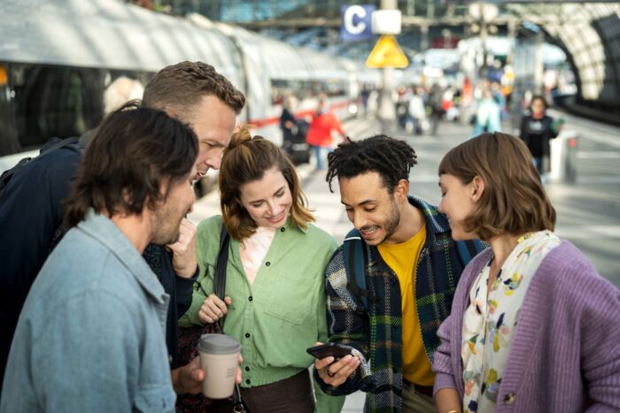 Mehrere Personen stehen am Bahnsteig zusammen und schauen auf ein Handy. Im Hintergrund ein ICE