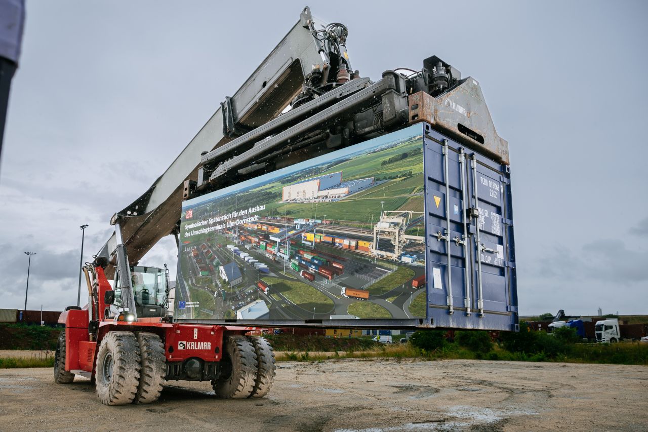 Ein Container ist zum Spatenstich des Umschlagterminals Ulm-Dornstadt mit einer bedruckten Plane geschmückt. Dieser wird von einem Greifstapler in der Luft gehalten.